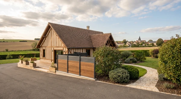 Photographie d'un paysage rural champêtre près de Compertrix, montrant une maison d'habitation avec un aménagement extérieur professionnel : terrasse, clôture moderne et jardin bien entretenu. La vue doit être prise depuis l'entrée ou l'accès à la propriété pour illustrer l'intervention d'un paysagiste, sans texte ni logo visible.